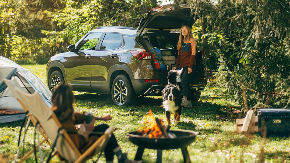 A Zeus Bronze Metallic Trailblazer ACTIV sits in a campsite surrounded by tall trees. Two people are in the campsite, along with various camping gear. A campfire burns in a small firepit in the center.