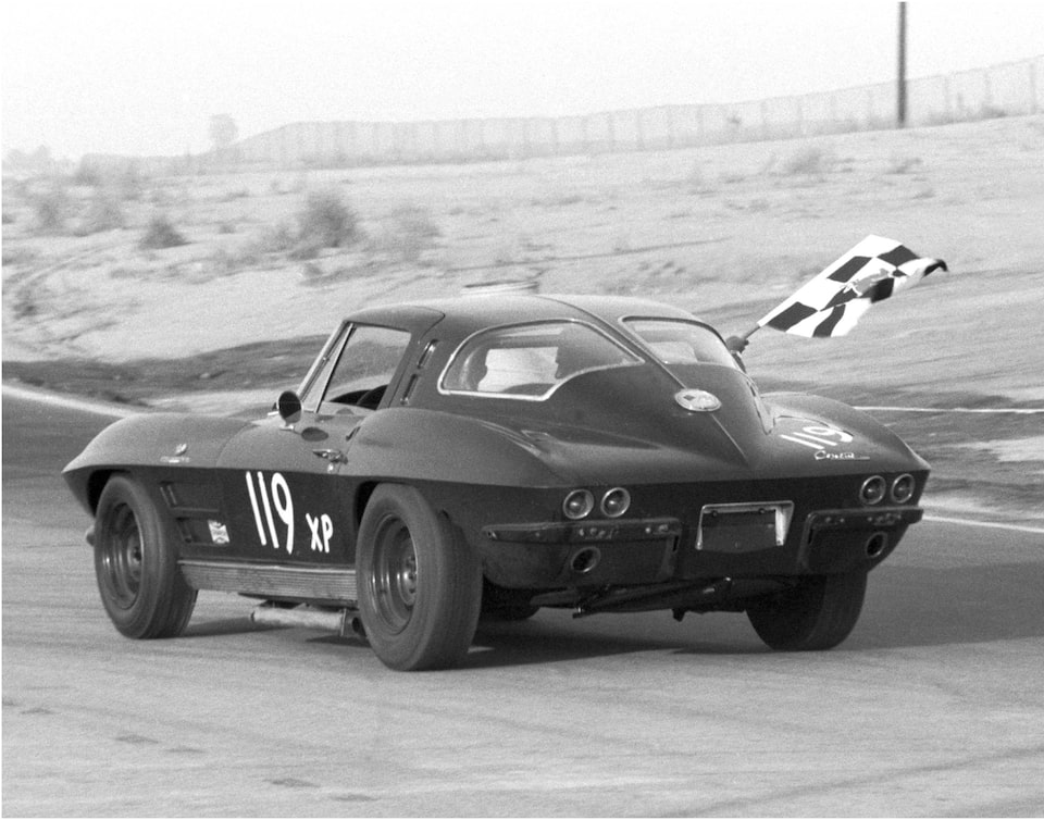 A historical photo of a 1963 split-window Corvette Sting Ray with the number 119 painted on the side and a black-and-white checkered flag waving out the passenger window driving on a roadcourse track.