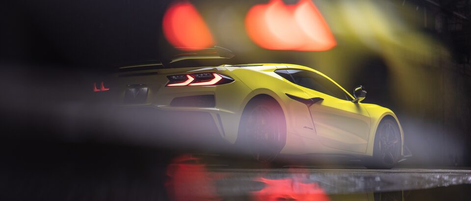 An Accelerate Yellow Metallic Corvette Z06 with tail lights lit sits in a dark area, with blurry red and yellow lights above that also reflect on the pavement under the car.