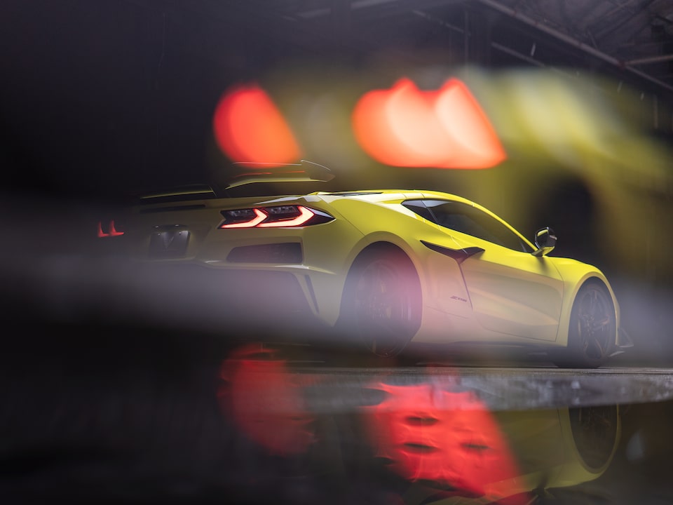 An Accelerate Yellow Metallic Corvette Z06 with tail lights lit sits in a dark area, with blurry red and yellow lights above that also reflect on the pavement under the car.
