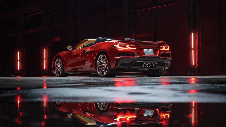 A red Corvette Z06 sits in a dark area, with red tail lights and red background lights glowing that also reflect on the wet pavement under the car.