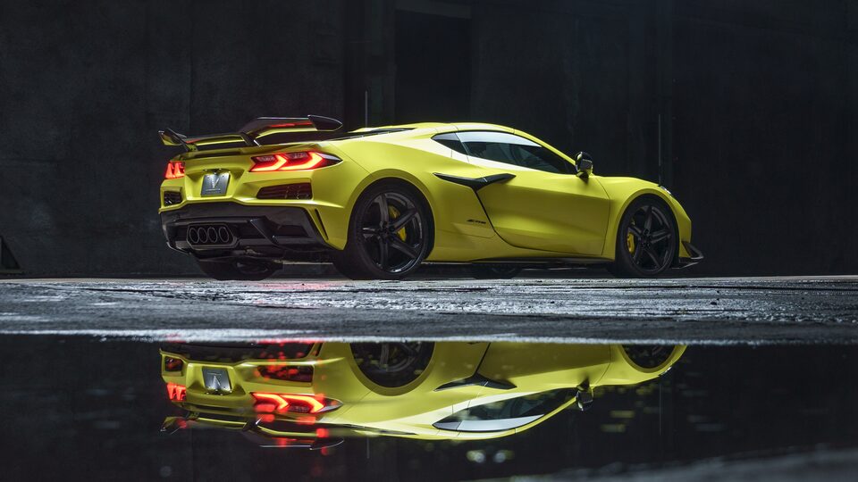 An Accelerate Yellow Metallic Corvette Z06 with tail lights lit sits in a dark area. The car is reflected in the wet pavement below.