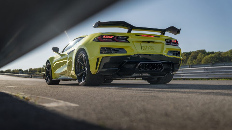 A yellow Corvette Z06 seen from behind the left rear sits on a track. Its distinctive Z07 Performance Package rear spoiler is outlined against the blue sky.
