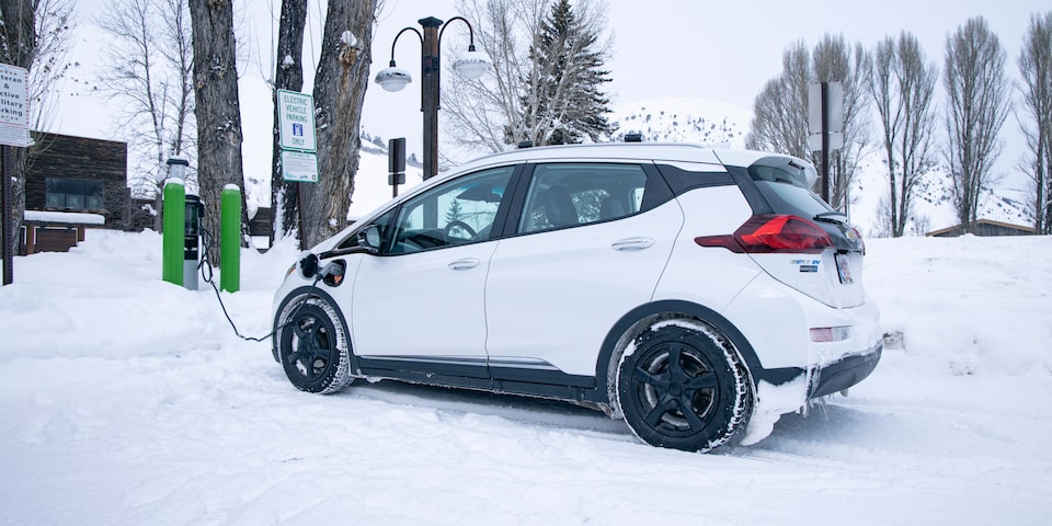 Un Bolt EV Blanco en un estacionamiento cubierto de nieve, enchufado a una estación de carga verde.