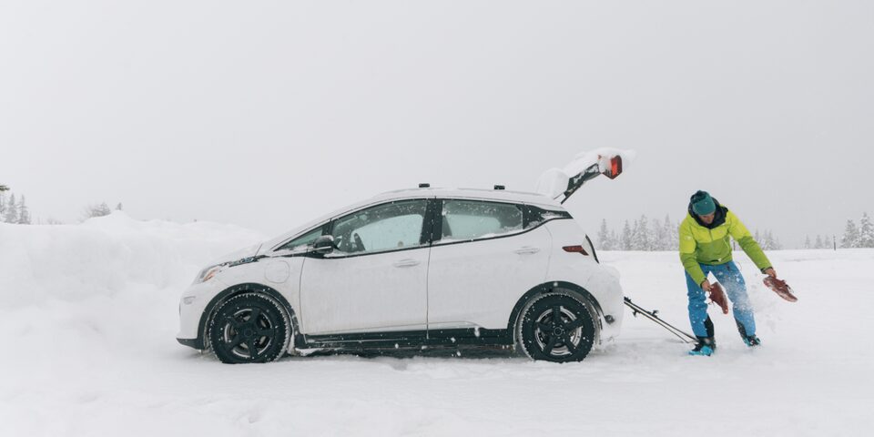 Greg Hill con una chaqueta verde neón y jeans azules carga sus equipos para esquiar en la cajuela abierta de un Bolt EV Blanco en un estacionamiento cubierto de nieve.