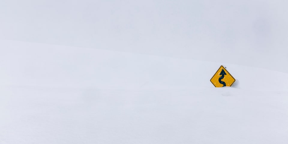 Una señal de advertencia amarilla que muestra un camino sinuoso al frente, asomando desde una pila de nieve.