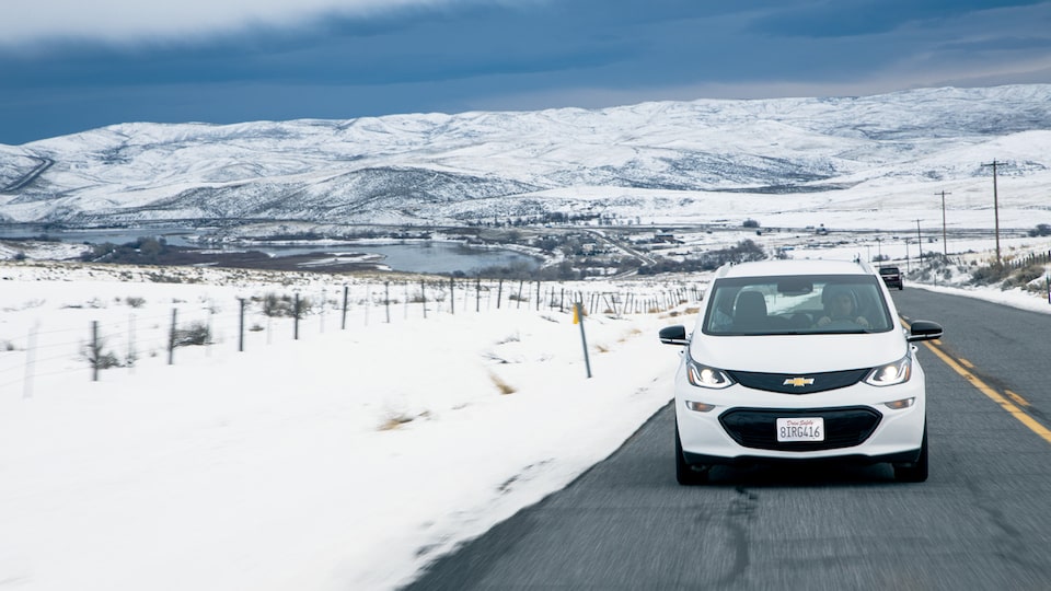 Un Bolt EV Blanco transitando un camino despejado rodeado de nieve en los bordes y colinas nevadas de fondo.