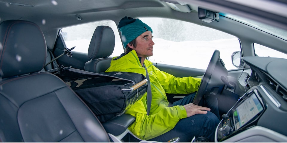 Greg Hill, con una chaqueta verde neón y una gorra turquesa, sentado en el asiento del conductor de un Bolt EV Blanco.