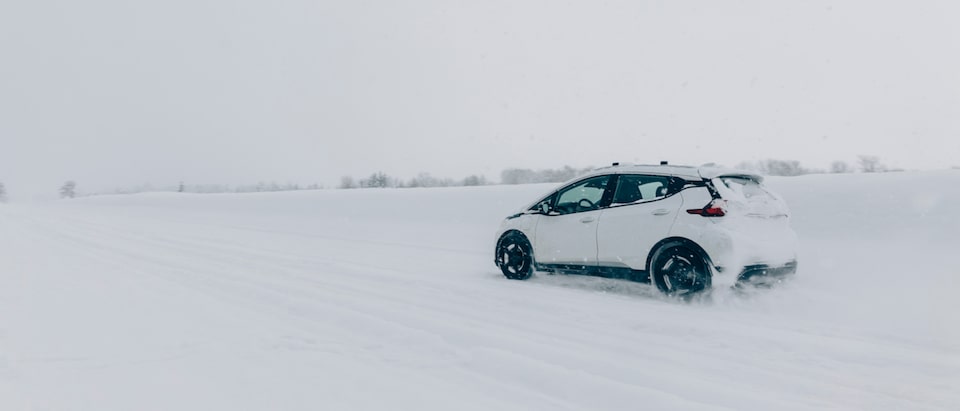 Un Chevrolet Bolt EV Blanco bajando por un camino plano cubierto de nieve con ningún otro vehículo a la vista y algunos árboles en la distancia.