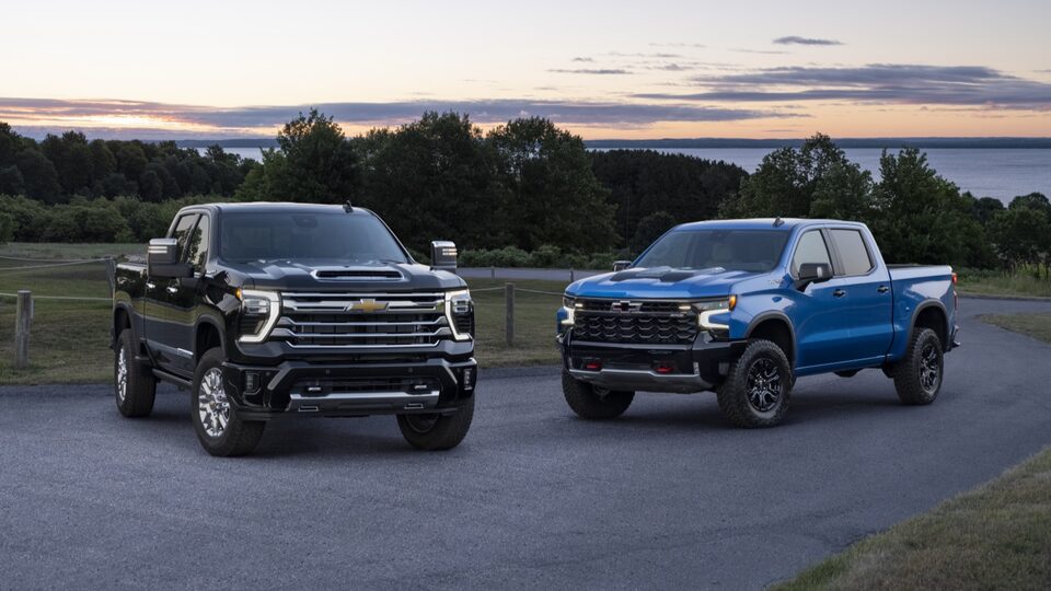 Two Silverado HD trucks sit next to each other on a narrow road with trees and a large lake in the background.