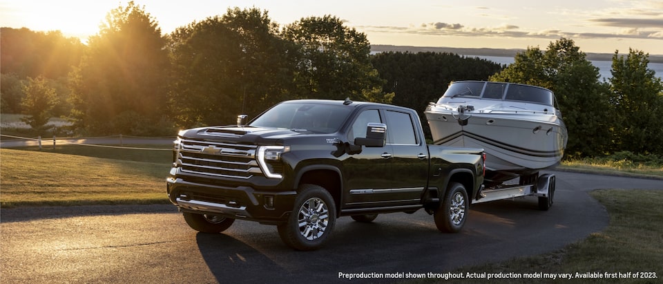 A black Silverado HD pulls a ski boat along a tree-lined road as the sun rises in the background.