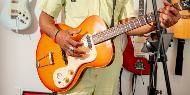 Redray Frazier toca la guitarra delante de un micrófono en su estudio. Otras guitarras cuelgan de la pared a sus espaldas.