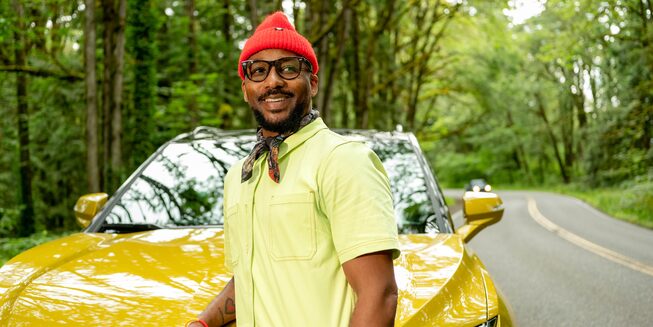 Redray Frazier, que tiene una camiseta amarilla y un sombrero rojo brillante, está parado delante de una Blazer de color amarillo brillante estacionada al costado de un camino rodeado de árboles.