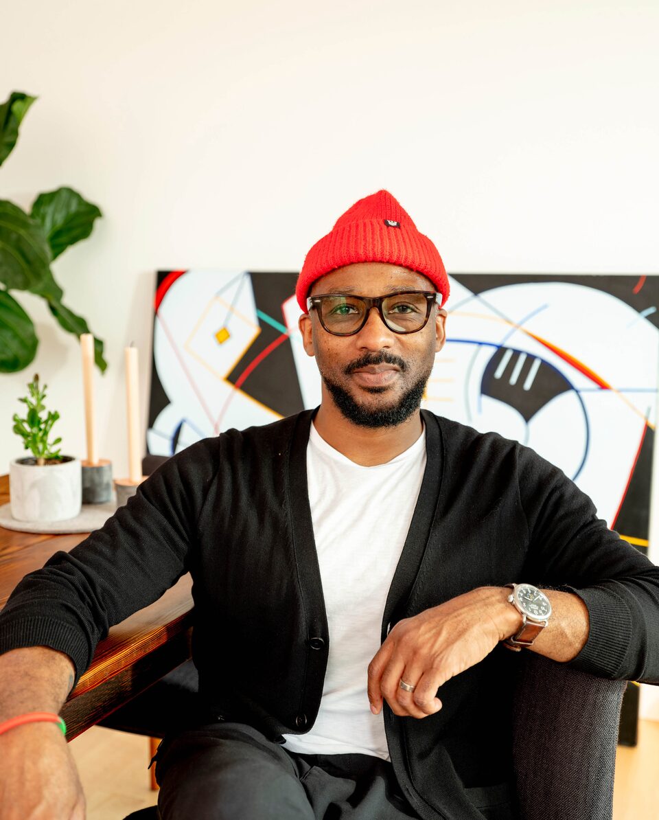 Redray Frazier, wearing a white t-shirt, black cardigan, and bright red hat, looks directly at the camera while sitting at a table in front of a large piece of art.