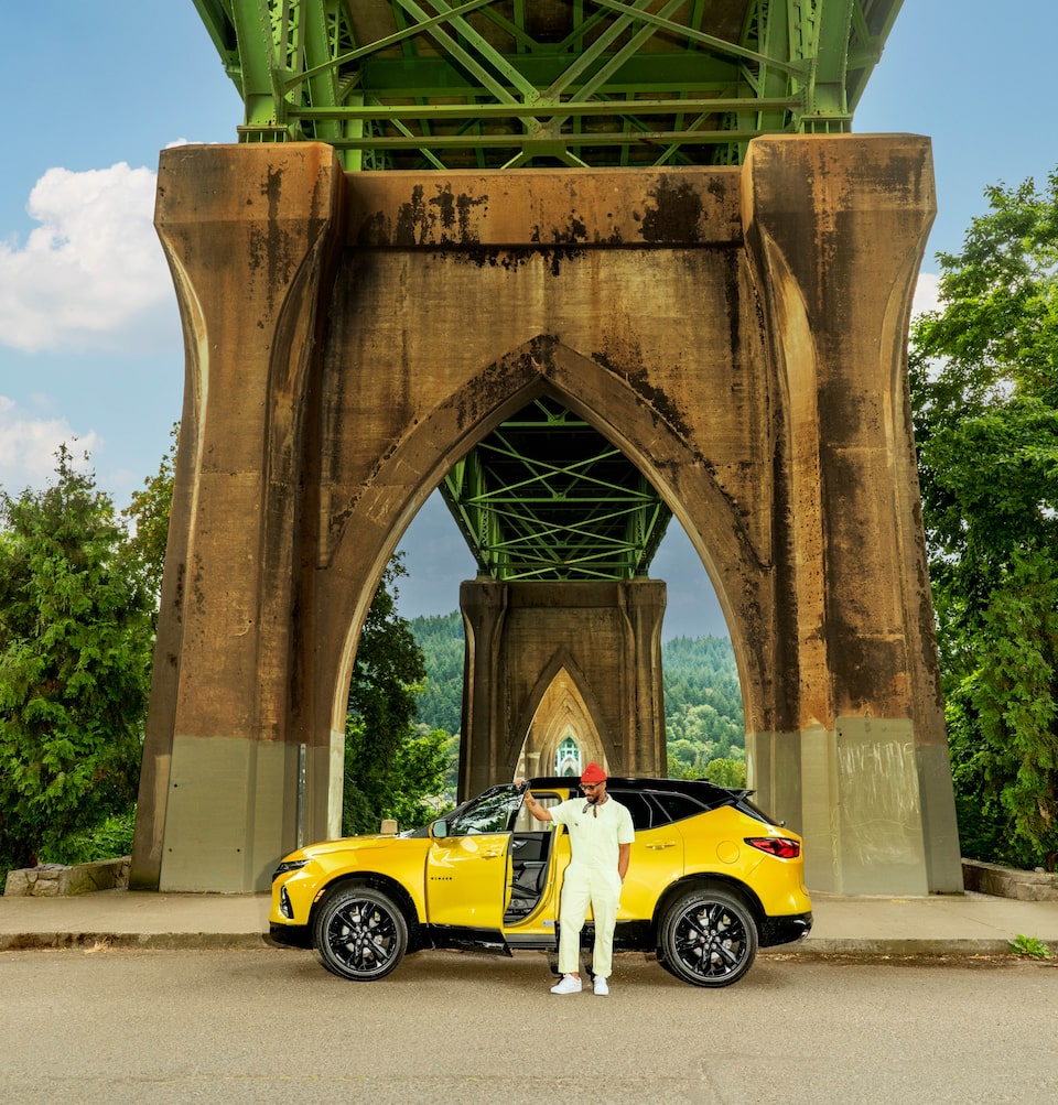 Redray Frazier, con una guitarra en la mano, se sienta en el techo de una Blazer color amarillo brillante aparcada bajo el puente St. Johns, en Portland (Oregón).