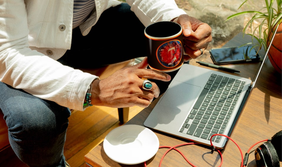 Un primer plano de las manos de Redray sosteniendo una taza de café colorida, sentado con una computadora portátil frente a una pequeña mesa.