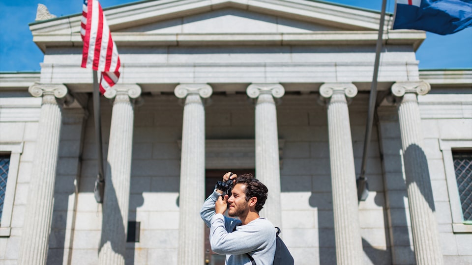 A man with a camera stands in front of the Bunker Hill Monument’s flags and columns.
