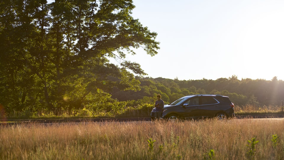 A man stands in front of an Equinox that is parked on the side of the road near a field.