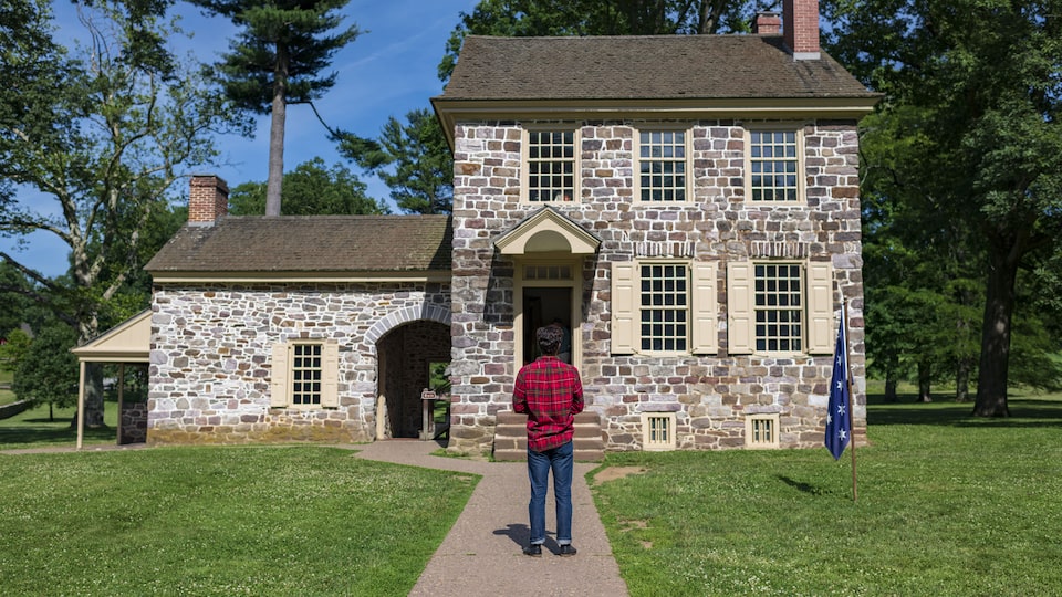 A man in a plaid shirt stands facing an old stone house.