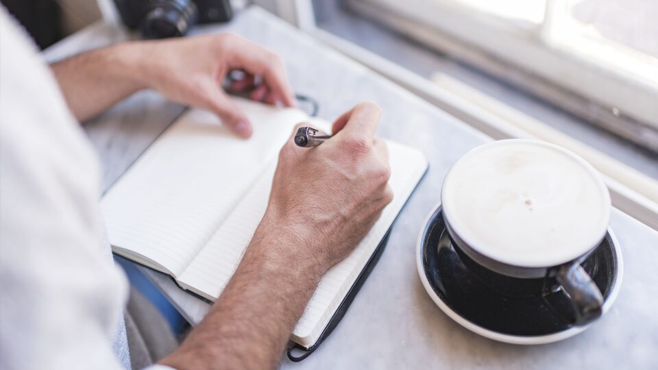 A close-up photo of a man’s hands as he writes in his notebook, with a coffee drink on the table.