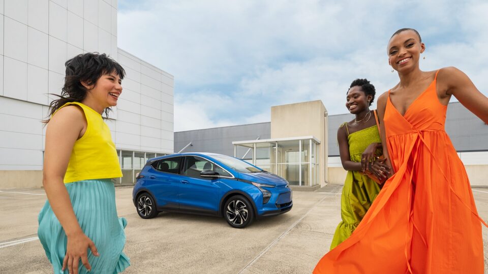 Tres personas con ropas coloridas posan cerca de una Bolt EUV azul estacionada en el nivel superior abierto de un estacionamiento.