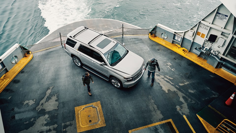 A father and son walking past a Chevy Tahoe SUV parked on the deck of a car ferry, seen from above and to the side.