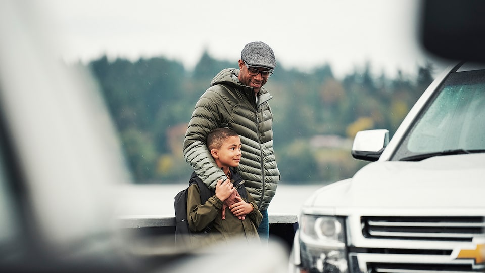 A father with his arm around his son stand on the deck of a car ferry next to a Chevy Tahoe SUV.