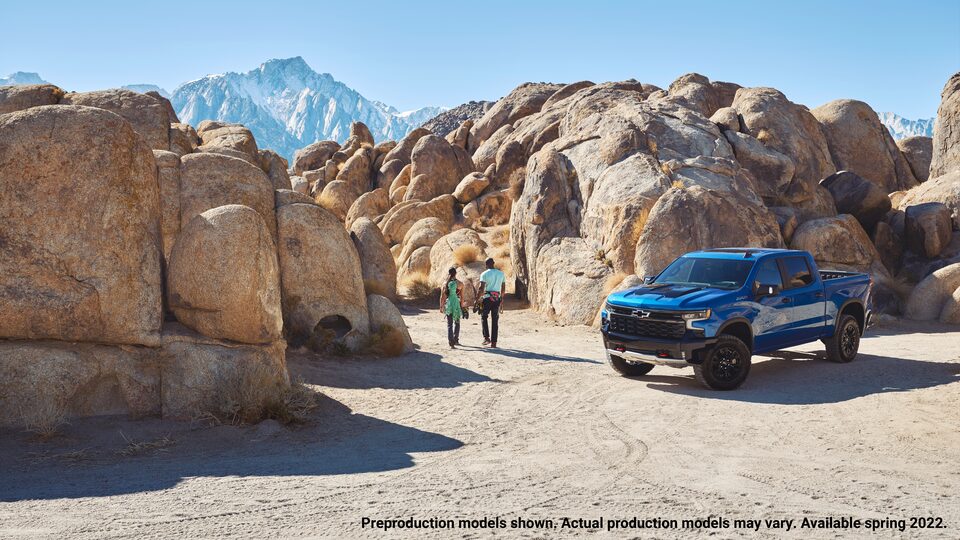 A blue Silverado ZR2 pickup sits on a flat gravel area with high, rounded boulders behind it and mountains in the background. Two people carrying climbing gear walk away from the truck toward the boulders.