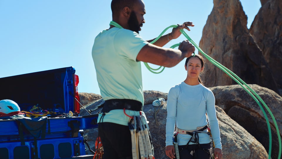 Two people stand behind the open tailgate of a blue Silverado ZR2. One of them is coiling a climbing rope around their arm.