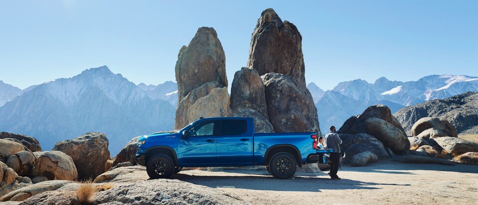 Una Silverado ZR2 azul estacionada al lado de un enorme saliente de roca, con montañas de fondo. Dos personas se sientan y paran sobre la puerta trasera abierta.