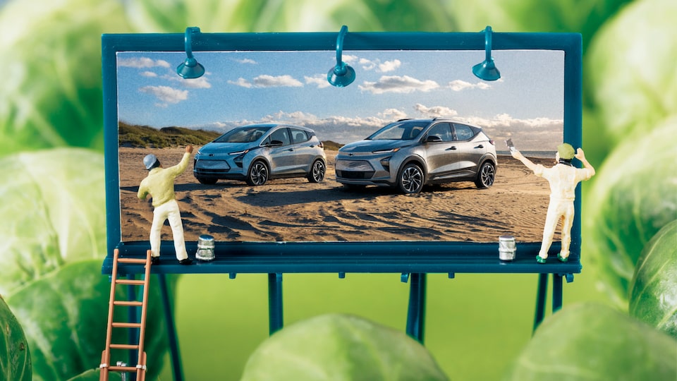 Una imagen tipo maqueta de una valla miniatura mostrando el Bolt EV y la Bolt EUV, con dos muñequitos pintándola, rodeada de coles de Bruselas para que parezcan árboles.