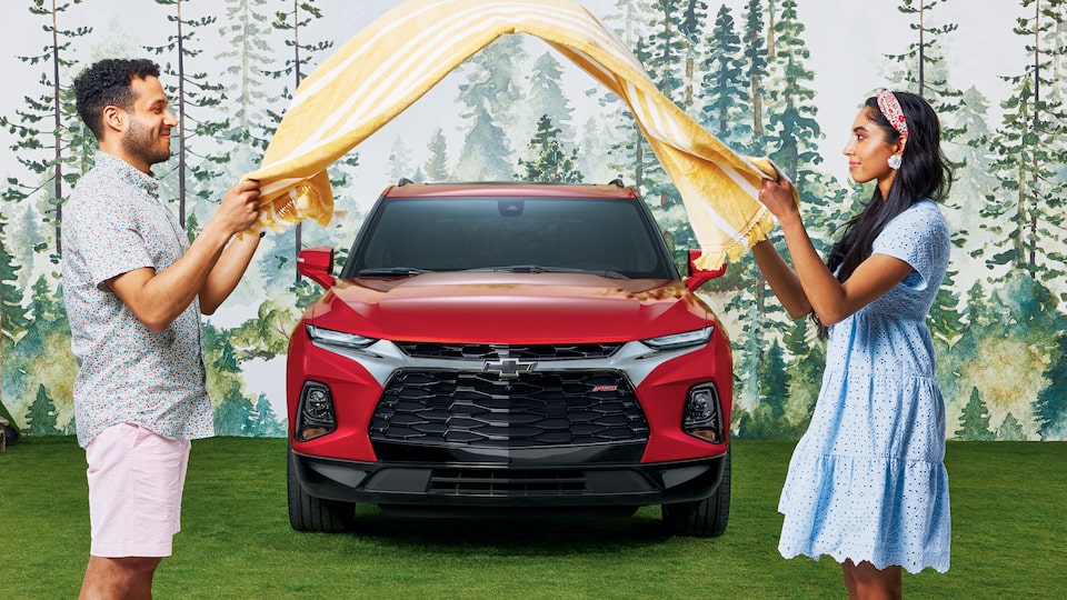 Dos personas, una frente a la otra, delante de una Chevy Blazer RS de color rojo sacuden una manta de pícnic con rayas amarillas entre las dos.