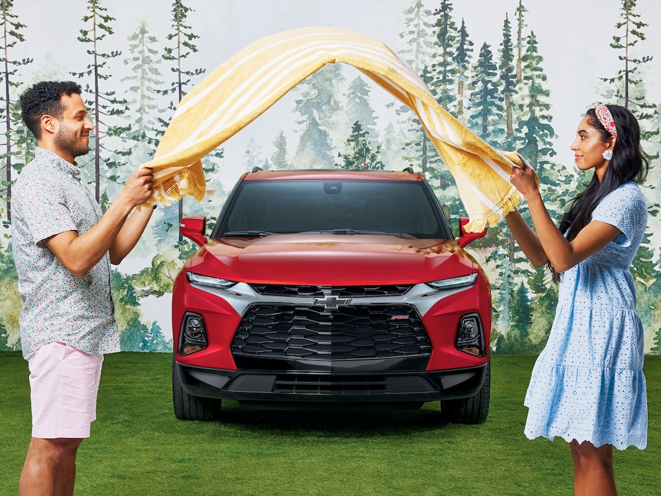 Dos personas, una frente a la otra, delante de una Chevy Blazer RS de color rojo sacuden una manta de pícnic con rayas amarillas entre las dos.