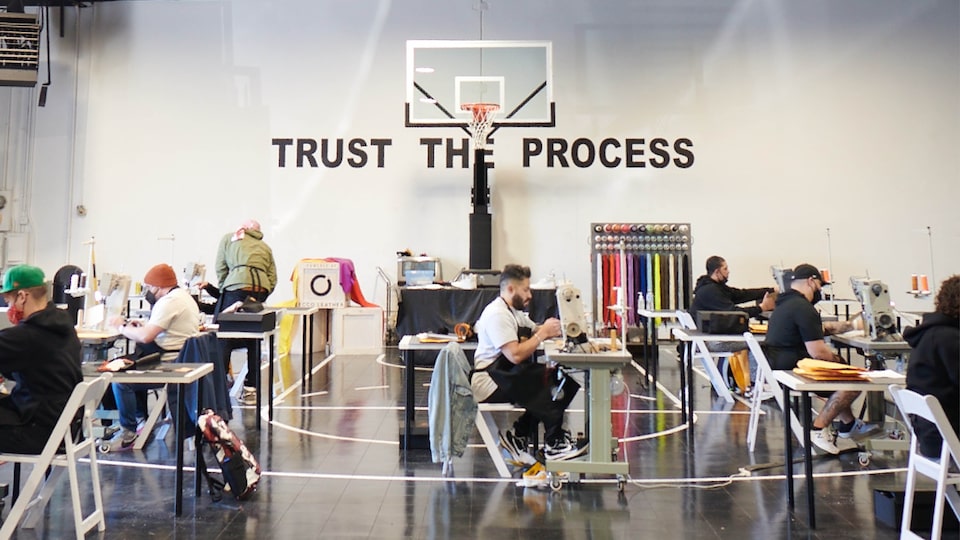 The Sneaker School se instaló en el gimnasio de una escuela, con casi una docena de personas sentadas en mesas con máquinas de coser. Un letrero detrás de una canasta de básquetbol que dice <em>Trust the Process</em> (Confía en el proceso).