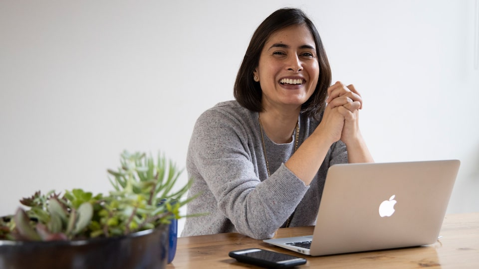 Una mujer sentada a una mesa sonriendo con una computadora portátil abierta y un teléfono celular frente a ella.