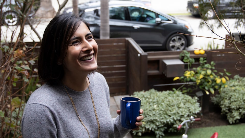 Una mujer sonriente está en el jardín sosteniendo una taza de café, con un Chevrolet Bolt EV color gris oscuro en la calle afuera del patio.