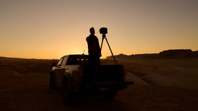 Silhouetted against the setting sun, a man stands in the bed of a Colorado ZR2 pickup with a large-format camera on a tripod.