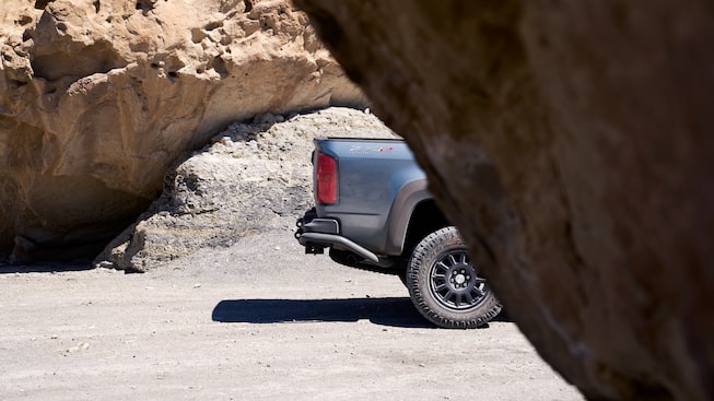 Just the rear of a gray Chevy Colorado ZR2 pickup is seen on a dirt road from behind a red rock formation.
