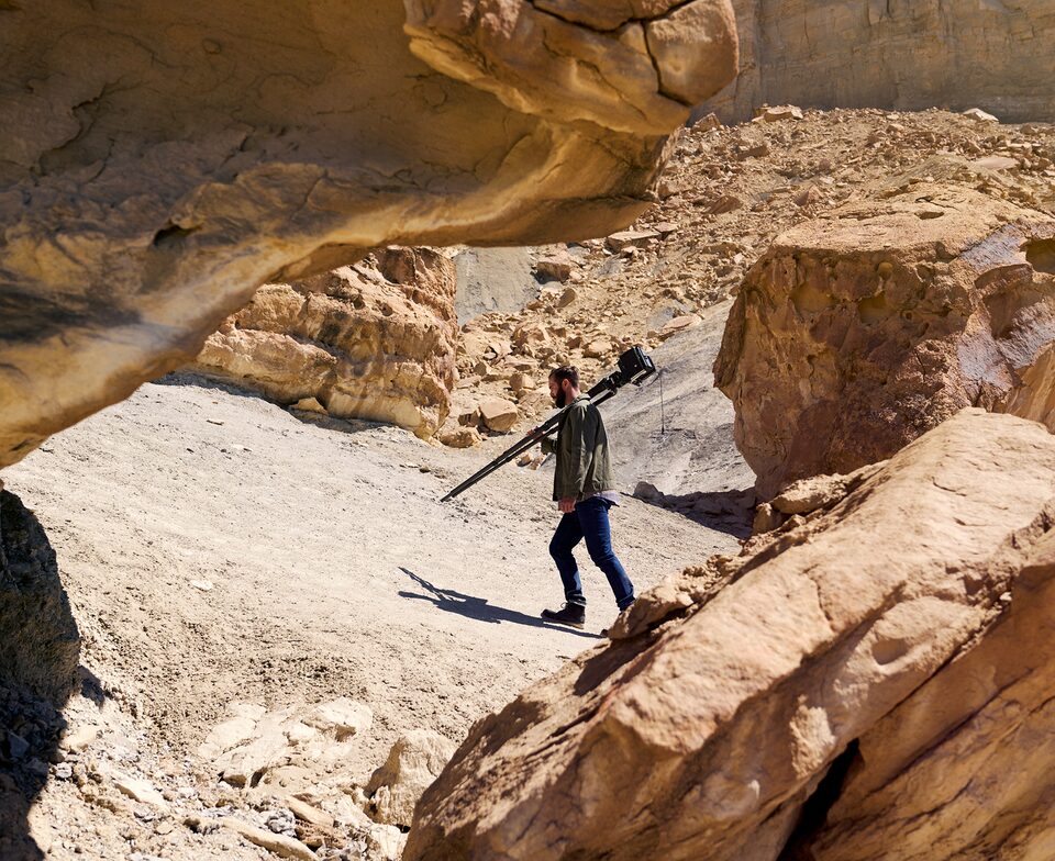 Hombre con una cámara de gran formato montada en un trípode plegable caminando por un sendero de tierra entre rocas rojas.