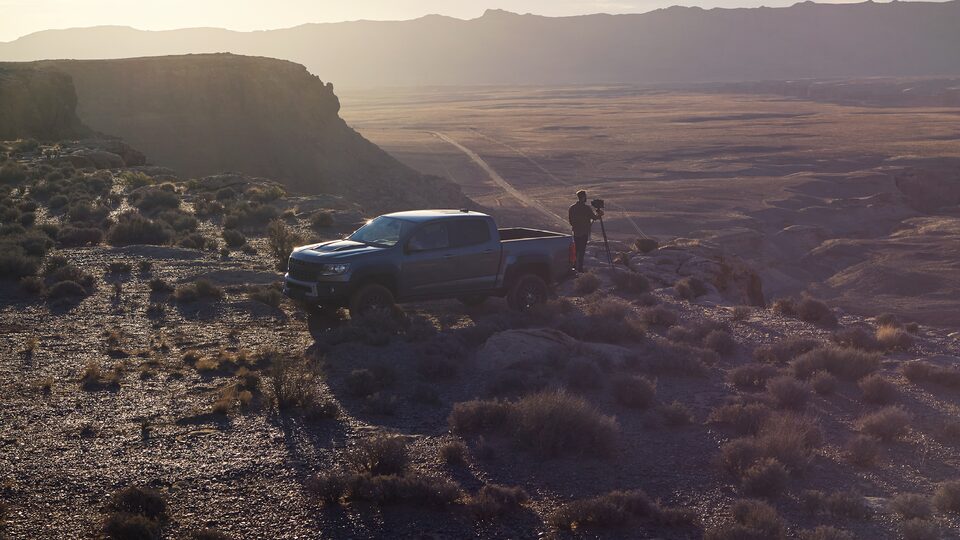 A gray Chevy Colorado ZR2 Bison sits on the edge of a steep, brush-covered hill at sunset. A man stands behind the truck using a large-format camera mounted on a tripod.