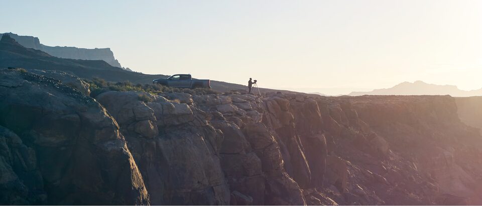 A wide shot of a cliff edge on the north rim of the Grand Canyon at sunset. Near the edge of the cliff are a gray Chevy Colorado ZR2 Bison pickup and a man standing at a camera mounted on a tripod.