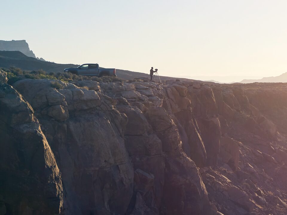 A wide shot of a cliff edge on the north rim of the Grand Canyon at sunset. Near the edge of the cliff are a gray Chevy Colorado ZR2 Bison pickup and a man standing at a camera mounted on a tripod.