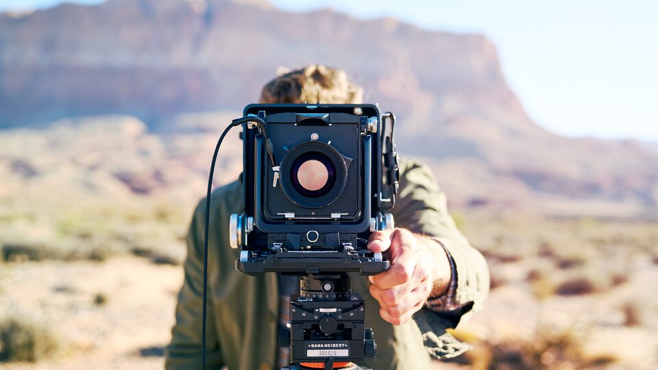 A person stands behind a large-format camera mounted on a tripod, with the Arizona desert and red rock cliffs in the distance.