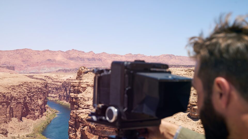 A man looks through the viewfinder of a large-format camera mounted on a tripod. The camera is aimed at the Colorado River flowing far below between red cliffs.