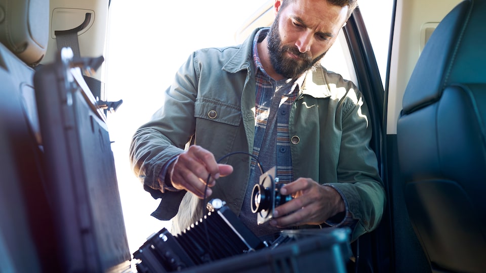 Seen from inside a Colorado ZR2 pickup, a man stands at the open back door assembling a large-format camera.