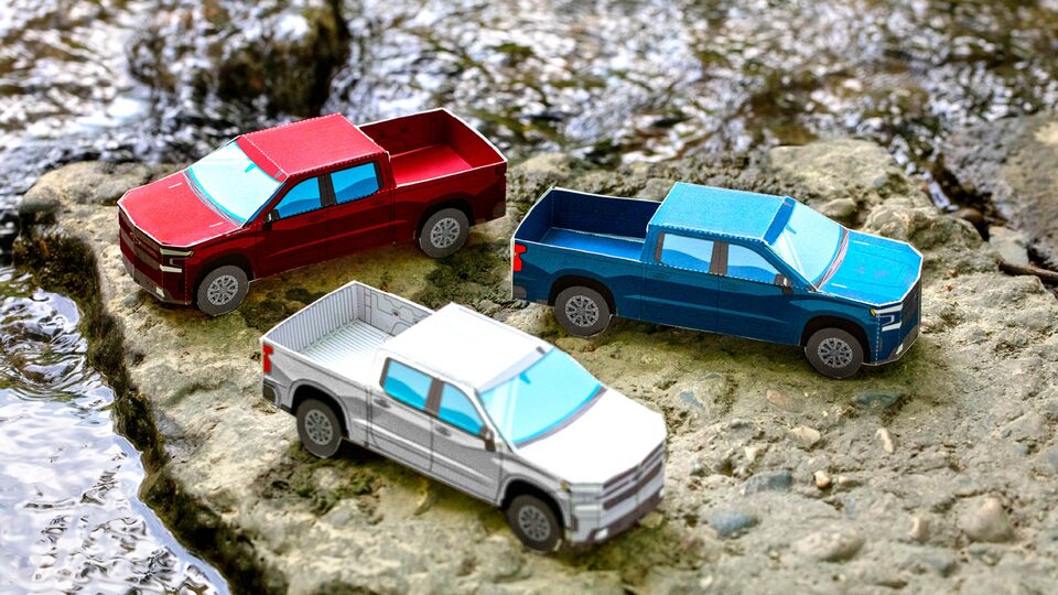 Miniature paper models of the 2020 Silverado RST Diesel in Cajun Red, Silver Ice Metallic, and Northsky Blue Metallic sitting on bare, rocky ground.