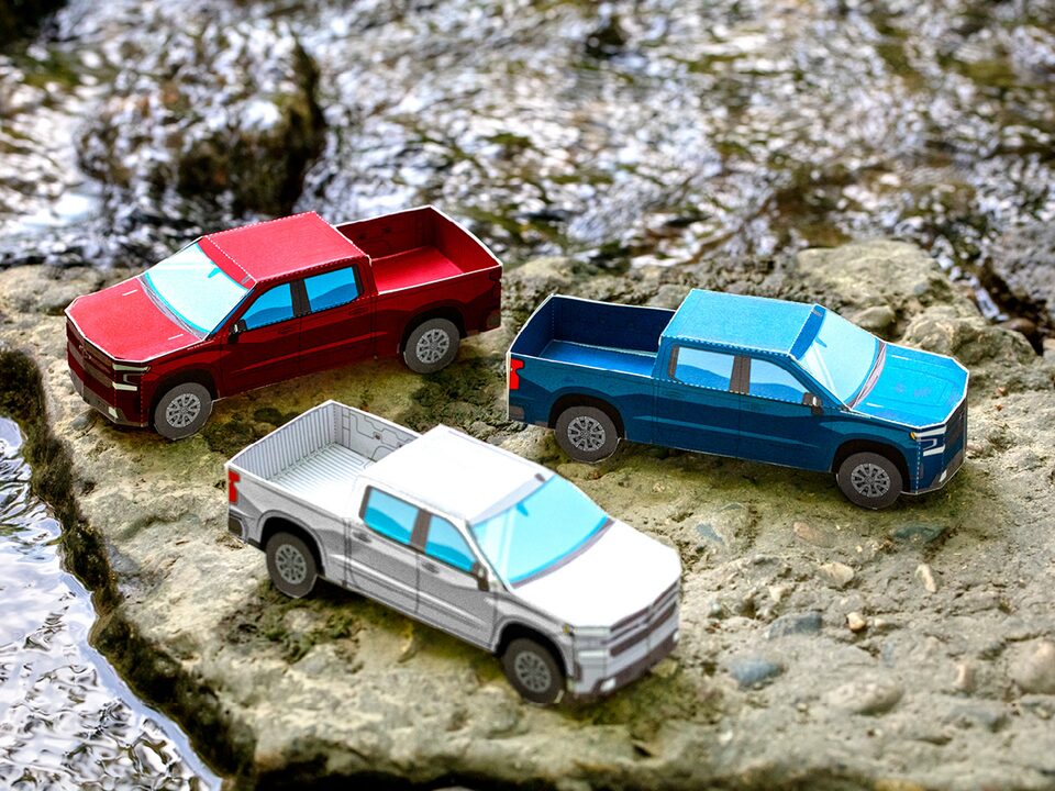 Miniature paper models of the 2020 Silverado RST Diesel in Cajun Red, Silver Ice Metallic, and Northsky Blue Metallic sitting on bare, rocky ground.