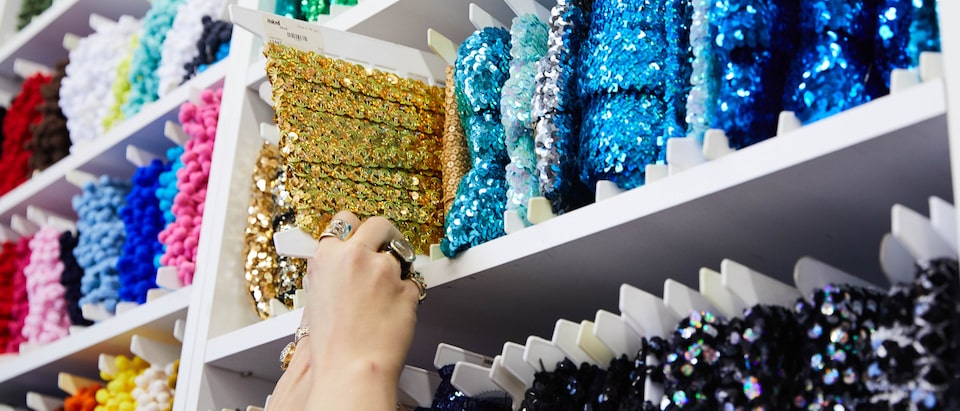 Shelves filled with colorful fabric trim. A person’s hands are reaching up to remove a spool of gold trim from a shelf.