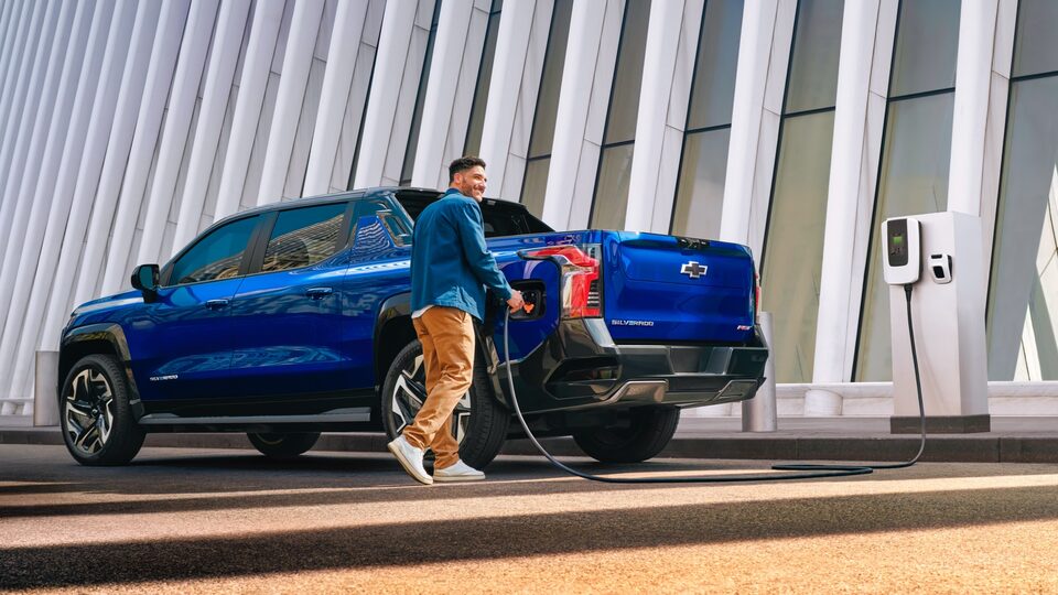 Una persona enchufa un cable de carga en una Silverado EV azul estacionada frente a un edificio de estilo contemporáneo.