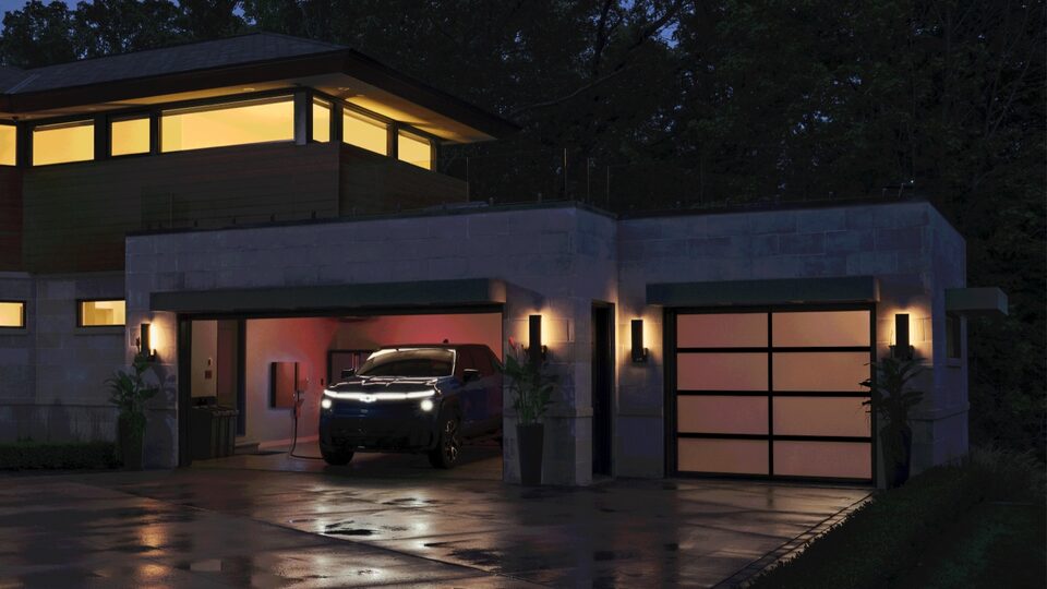 Una Silverado EV enchufada en un cargador en el interior del garaje de una casa moderna iluminadas por luces en el interior y exterior, vista de noche a través de la puerta abierta del garaje.